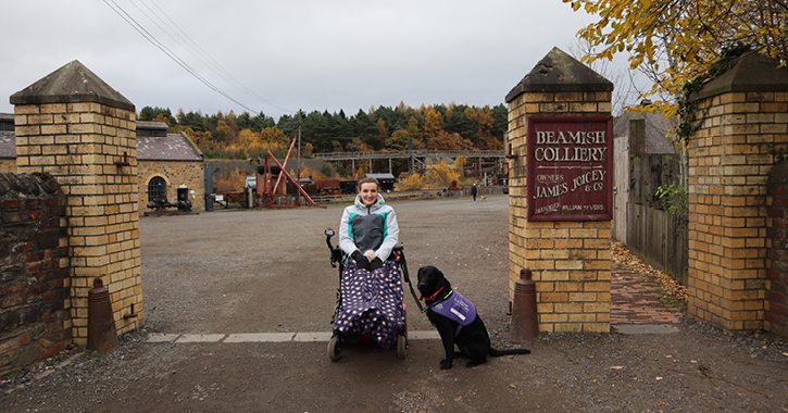 woman sat in wheelchair with assistance dog by her side at the entrance to 1900s colliery at Beamish Museum